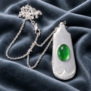 May include: A silver pendant necklace featuring a smooth, elongated, shield-shaped silver plate with a polished finish. A single, oval-shaped, vibrant green cabochon stone is set in the centre. The pendant hangs from a delicate silver chain.