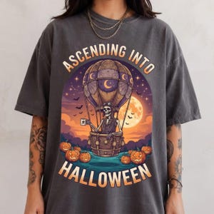 May include: A charcoal grey t-shirt featuring a graphic of a skeleton in a hot air balloon, holding a mug. The design includes a full moon, bats, and pumpkins on the ground. The text reads ASCENDING INTO HALLOWEEN in orange lettering.