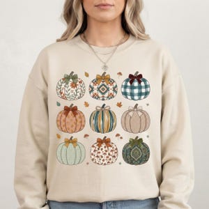 May include: A beige crewneck sweatshirt featuring a grid of nine illustrated pumpkins. Each pumpkin displays a unique pattern, including floral, geometric, plaid, striped, cable knit, and paisley designs, topped with small bows and scattered leaves.