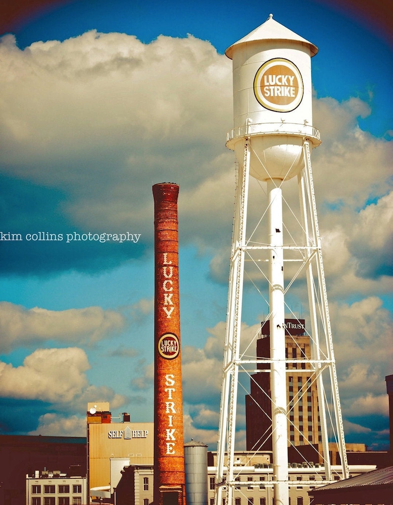 May include: A red brick chimney with the words "Lucky Strike" painted on it stands next to a white water tower with the same words on it. The water tower is a lattice structure with a large white tank at the top. The sky is blue with white clouds.