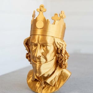 May include: A metallic gold-coloured bust of a person with long, wavy hair wearing a crown with circular and cross-shaped details. The figure has a defined facial structure, a collared shirt, and a smooth, reflective finish across the entire surface.