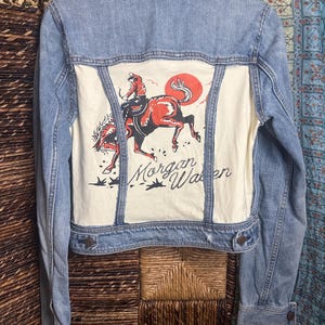 May include: A light blue denim jacket featuring a cream-colored back panel with a graphic print of a person riding a bucking horse under a red sun. The text Morgan Wallen is printed in black script below the horse illustration.