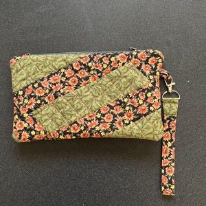 May include: A rectangular quilted wristlet pouch featuring a diagonal patchwork pattern of black fabric with orange and yellow floral prints alternating with olive green fabric with a fern leaf pattern, including a matching detachable wrist strap.