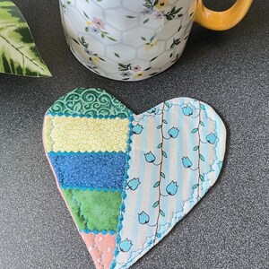 May include: A heart-shaped fabric coaster featuring a patchwork design of green, yellow, blue, and pink patterned fabric on one side and a light blue and white striped fabric with a blue tulip vine print on the other, with visible stitching.