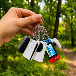 May include: A metal keyring featuring multiple miniature gaming console and controller charms, including a white console with a black circular vent, a black console, a handheld device with a blue and red controller, and a white game controller.
