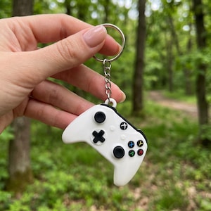 May include: A white video game controller keychain featuring a black directional pad, two black analog sticks, and four colored action buttons labeled A, B, X, and Y. The controller is attached to a silver metal split ring by a short chain.