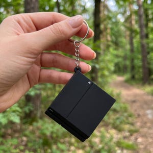 Può includere: Un portachiavi quadrato nero a forma di console di gioco, dotato di anello e catena in metallo argentato. La superficie nera opaca presenta un piccolo logo PlayStation bianco nell'angolo in alto a destra e la scritta Sony sul bordo sinistro.