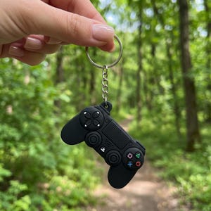May include: A black silicone video game controller keyring featuring a metal split ring and chain. The controller design includes a directional pad, two analogue sticks, a central touchpad, and four action buttons with green, pink, blue, and red symbols.
