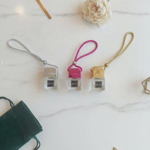 May include: Three small glass car air freshener bottles with square bases and metallic caps in silver, hot pink, and gold. Each bottle features a black label with the word Grid in white text and includes a matching colored hanging cord loop.