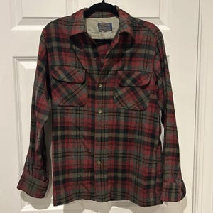 May include: A long-sleeve button-down shirt featuring a plaid pattern in shades of red, black, and olive green. The shirt includes a pointed collar, two chest pockets with buttoned flaps, and a Pendleton brand label inside the neck area.