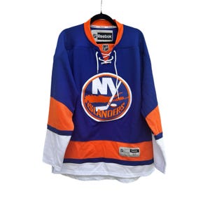 May include: A blue, orange, and white Reebok New York Islanders hockey jersey featuring a lace-up collar, the team logo on the chest, and an NHL shield. The jersey includes a size M tag on the lower front and Reebok branding on the inner neck label.