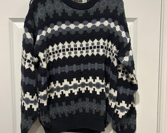 May include: A long-sleeve crew neck sweater featuring a repeating geometric pattern in shades of black, charcoal gray, light gray, and off-white. The knit garment has a ribbed collar, cuffs, and hem, with a visible brand label at the inner neckline.