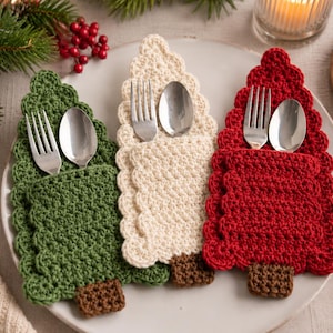 May include: Three crochet Christmas tree-shaped cutlery holders in green, cream, and red. Each holder features a small brown square base and a pocket for a silver fork and spoon. The edges of the trees have a scalloped texture, and they are arranged on a plate.