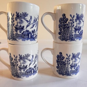 Può includere: Quattro tazze in ceramica bianca disposte in due pile, ciascuna caratterizzata da un motivo a salice blu. Il design raffigura scene tradizionali con pagode, barche sull'acqua e uccelli in volo. Ogni tazza ha un manico curvo.