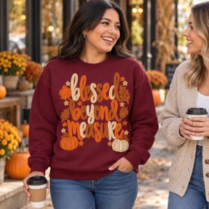 May include: A maroon crewneck sweatshirt featuring the text "blessed beyond measure" in a stylized orange, yellow, and cream font. The graphic includes illustrations of autumn leaves, small stars, an acorn, and two pumpkins.
