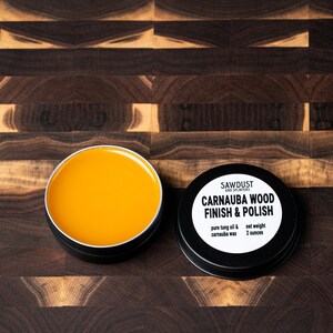 May include: A small black tin with a white label that reads "SAWDUST CARNAUBA WOOD FINISH & POLISH pure tung oil & carnauba wax net weight 2 ounces". The tin is open and the yellow wax inside is visible.