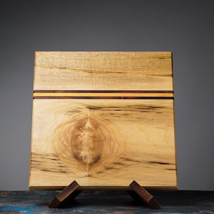 May include: A rectangular wooden cutting board with a light brown base and a decorative stripe of dark brown and yellow wood. The board is supported by two small wooden stands. The wood grain is visible throughout the board.