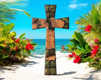 May include: A wooden cross with a unique design stands on a sandy beach with a tropical backdrop. The cross features a combination of brown wood and green resin, with a natural wood grain pattern. Red flowers and palm trees frame the cross against a blue sky and ocean.