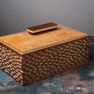 May include: A rectangular wooden box with a light-colored top and a dark-colored, patterned body. The lid has a small, rectangular handle. The box is made of wood and has a clear finish. The box is likely used for storage.