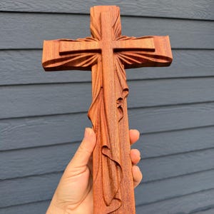 May include: A reddish-brown wooden cross featuring intricate carved details that resemble draped fabric flowing down the vertical beam and radiating from the center intersection, showcasing a smooth, polished grain texture throughout the piece.