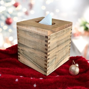 May include: A square wooden tissue box with a light brown wood grain pattern. The box has a small opening on the top for tissues. The box is sitting on a red blanket with white snowflakes.