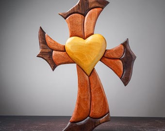 May include: A wooden cross with a heart-shaped center. The cross is made of various wood tones, including light brown, dark brown, and orange. The heart is a light yellow color. The cross has a decorative, stylized design.