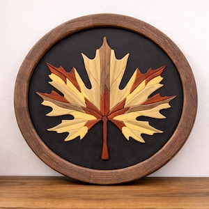 May include: A round wooden wall art piece featuring a maple leaf design. The leaf is crafted from various wood tones, including brown, yellow, and red, set against a black background within a brown frame. Decorative wall art.