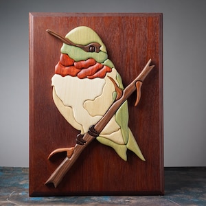 May include: Wooden hummingbird art piece mounted on a rectangular, dark brown wooden board. The hummingbird is crafted from various wood tones, including light green, red, and cream. It is perched on a brown branch, creating a nature-themed wall decor.