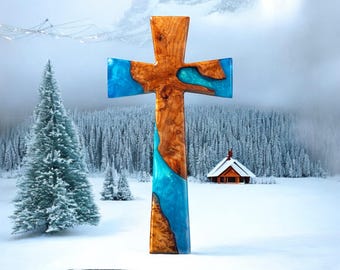 May include: A wooden cross with a blue and brown epoxy resin inlay. The cross is standing on a snowy surface with a small cabin in the background.