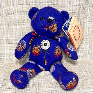 May include: A blue plush teddy bear featuring a repeating sun-like pattern in orange, yellow, and white. The bear wears a black cord necklace with a silver-toned circular pendant and has a paper tag attached to its ear labeled PENDLETON.