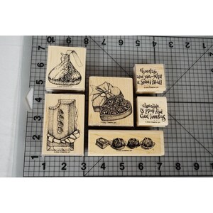 Op de afbeelding: Een set van vijf houten rubberstempels met chocolade-illustraties en tekst. De stempels bevatten een chocoladekus, een doos bonbons, een chocoladereep en twee tekststempels met Chocolate and you-what a sweet treat! en Chocolate is proof that God loves us. De stempels liggen op een snijmat met centimeter-markeringen.