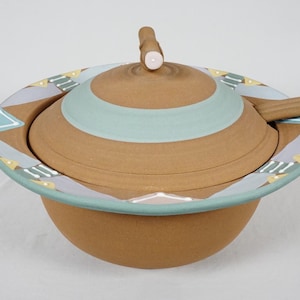 Può includere: Una ciotola in ceramica color terracotta con coperchio abbinato e una piccola maniglia. Il bordo presenta un motivo decorativo di forme geometriche azzurre, bianche e gialle. Un piccolo accento circolare chiaro si trova sulla maniglia del coperchio.