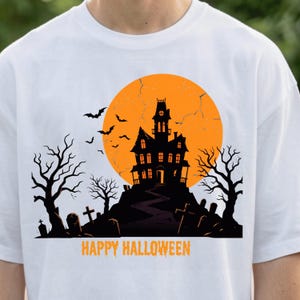 May include: A white t-shirt featuring a graphic of a black haunted house silhouette set against a large orange moon. The design includes bare trees, tombstones, flying bats, and the orange text "HAPPY HALLOWEEN" printed at the bottom.