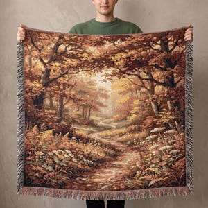 Könnte beinhalten: Eine gewebte Wohndecke mit einer detaillierten Herbstwaldszene, einem gewundenen Waldweg, goldenem und braunem Laub, hohen Bäumen und Wildblumen. Das rechteckige Textil hat an der Ober- und Unterseite einen dekorativen Rand mit Fransen.