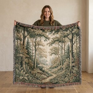 May include: A woven throw blanket featuring a detailed forest landscape scene with tall trees, dense foliage, and a winding path. The tapestry-style design uses shades of green, brown, and cream, finished with a decorative fringed edge on all sides.