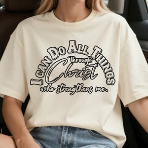 Puede incluir: Una camiseta de manga corta color crema con texto en negro que dice I CAN DO ALL THINGS Through Christ who strengthens me. El diseño utiliza una mezcla de letras de bloque en negrita y una fuente cursiva centrada en la parte delantera.