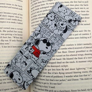 May include: A rectangular bookmark featuring a black and white comic strip pattern of the character Snoopy. The design includes multiple panels with Snoopy in various poses, with one instance of the character wearing a bright red sweater.