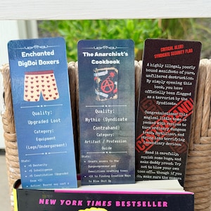 May include: Three rectangular bookmarks with game-themed text and graphics. The left is blue with heart-patterned boxers, the center is dark blue with a bomb icon, and the right is dark red with black text about a syndicate security flag.