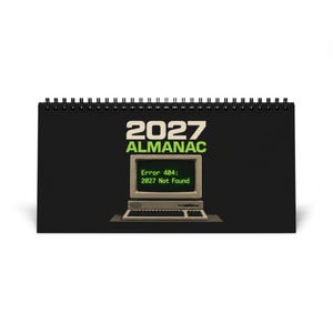 Può includere: Un calendario da scrivania con rilegatura a spirale nera che riporta il testo 2027 ALMANAC in bianco e verde. Sotto il testo c'è l'illustrazione di un computer beige vintage con un monitor che mostra il testo verde Error 404: 2027 Not Found.