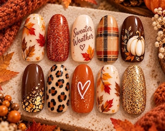 May include: A set of ten press-on nails featuring autumn-themed designs including red and orange maple leaves, copper glitter, a cream nail with the text Sweater Weather, a brown plaid pattern, a white pumpkin, leopard print, and a heart.