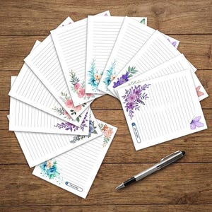 May include: A collection of white lined stationery sheets arranged in a fan shape, each featuring watercolor floral illustrations in shades of pink, blue, and purple, accompanied by a silver ballpoint pen with a black grip and a silver clip.
