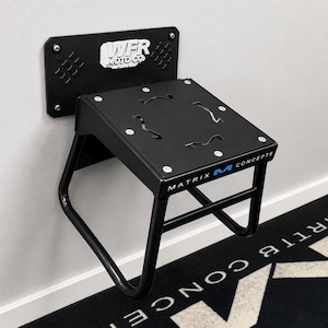 May include: A black wall-mounted motorcycle stand featuring a rectangular base with the text WFR MOTO CO. and a top platform labelled MATRIX CONCEPTS. The stand has a tubular metal frame and a flat surface with four cut-out details secured by screws.