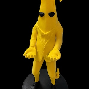 May include: A yellow banana-shaped figure wearing a suit, bowtie, and black shoes. The figure has black oval eyes, a textured surface, and is standing with arms outstretched, palms facing upward. A small object is attached to the figure's left leg.