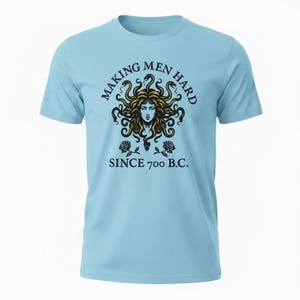 Puede incluir: Una camiseta azul claro con un gráfico de Medusa con cabello de serpientes doradas, flanqueada por dos rosas negras. El texto "MAKING MEN HARD" aparece arriba y "SINCE 700 B.C." abajo, impreso en una fuente oscura y negrita con serifa.