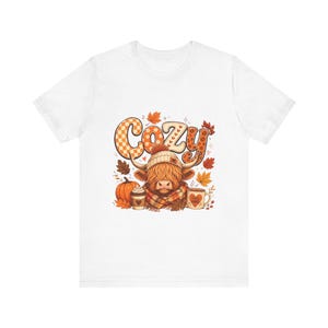 May include: A white short-sleeve t-shirt featuring a graphic of a Highland cow wearing a knit hat and scarf. The cow is surrounded by autumn leaves, a pumpkin, a coffee cup, and a mug labeled Fall Vibes. The word Cozy is printed above the cow.