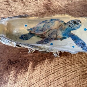 May include: An oyster shell featuring a detailed illustration of a blue and white sea turtle. The interior of the shell is painted cream and decorated with small blue dots and two white pearls at the left edge. It rests on a small silver stand.