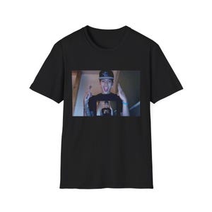Puede incluir: Una camiseta negra de manga corta con un estampado gráfico rectangular centrado. El estampado muestra a una persona con gorra de béisbol oscura, sonriendo con la boca abierta y haciendo gestos con las manos, con una versión más pequeña.