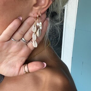 Puede incluir: Un conjunto de tres pendientes dorados con conchas cauri blancas naturales y una pequeña perla blanca. El set incluye un pendiente largo con tres conchas apiladas y dos aros más pequeños con detalles de concha y perla en cada uno de ellos.
