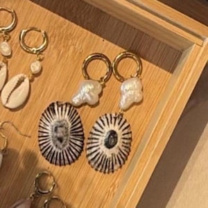 Puede incluir: Un par de pendientes de aro en tono dorado con perlas barrocas blancas colgantes y conchas ovaladas con un distintivo patrón de rayas radiales en blanco y negro. Los pendientes cuentan con un mecanismo de cierre seguro para el lóbulo de la oreja.
