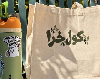 May include: A light green Owala water bottle with an orange lid and a sticker that reads Mentally in Lebanon, a beige canvas tote bag with dark green Arabic text and olive branch graphics, and a light green smartphone case with a triple camera lens.